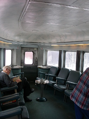 [Windows all around and on the back door provide lots of light into the seating area. Seats with armrests line both sides of the car. A man sits and reads in one on the left.]